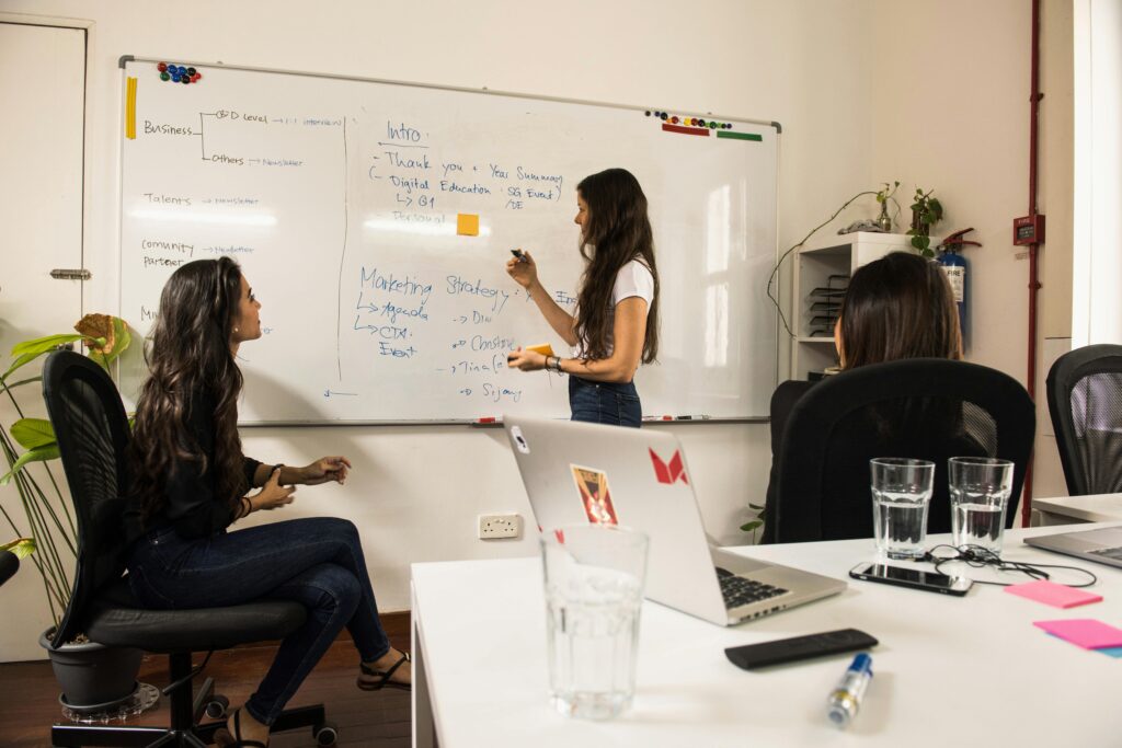 Coaching y mentoring profesional Business women discussing strategy at a whiteboard in a modern office setting.